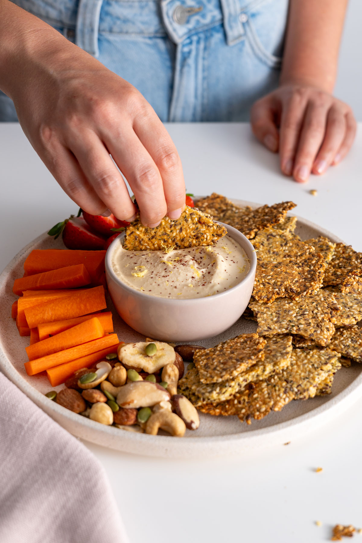 Sarah Cobacho dipping a healthy seeded cracker into creamy dip with a colorful platter of strawberries, carrot sticks, and nuts.