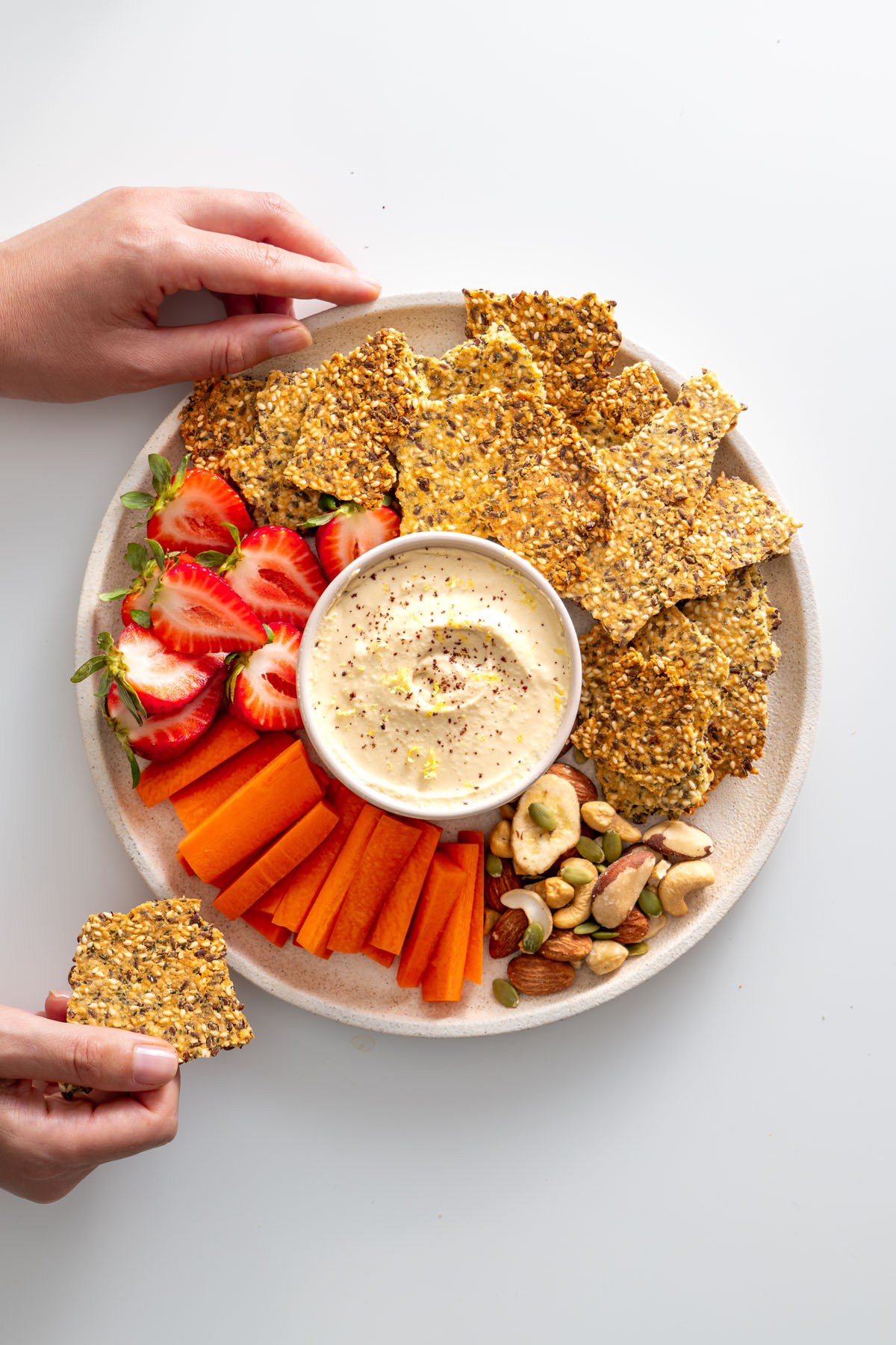 Overhead shot of a vibrant platter featuring healthy seeded crackers, fresh strawberries, carrot sticks, nuts, and creamy dip.