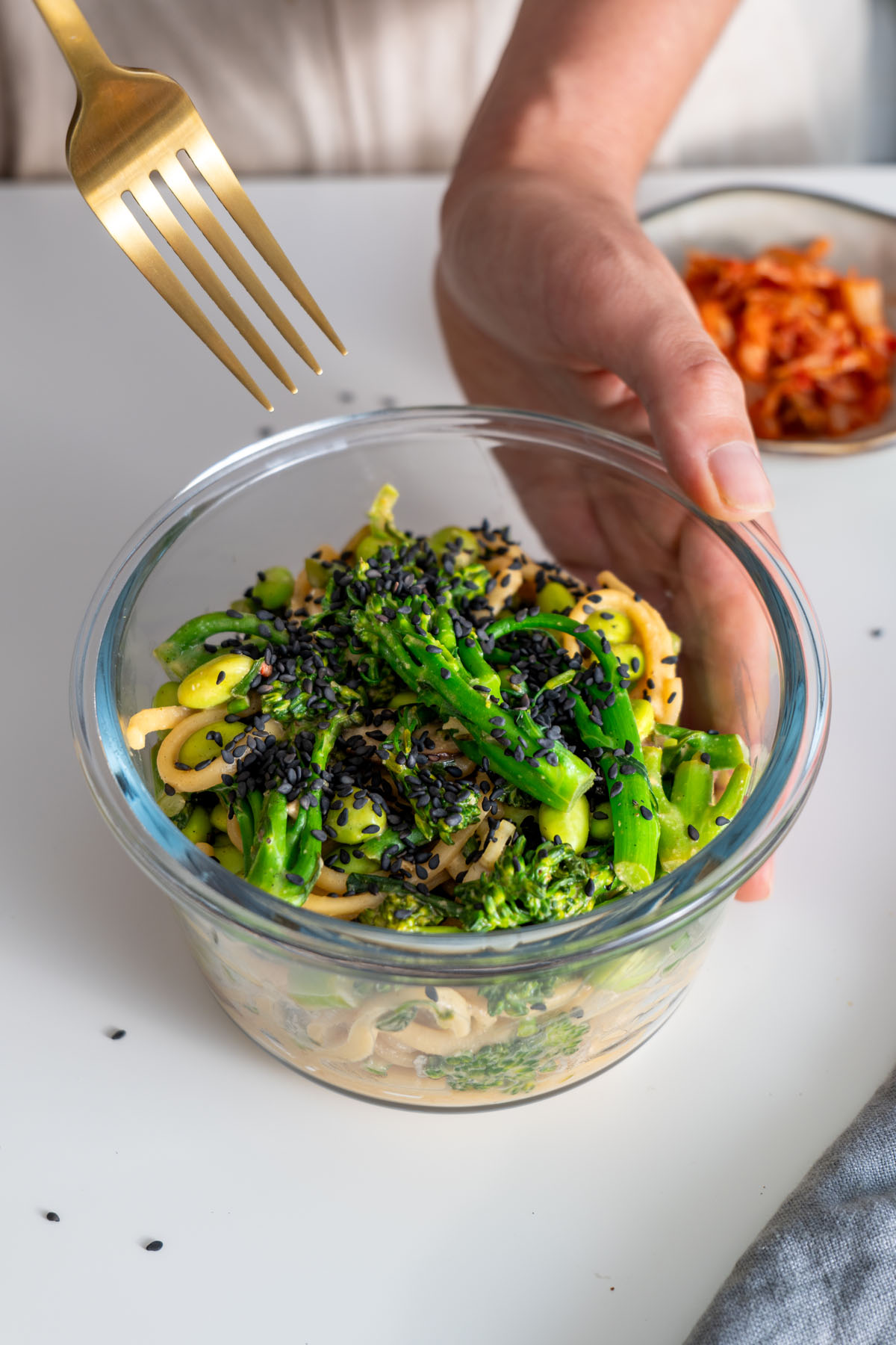 Close-up of a glass container with high-protein udon noodles topped with black sesame seeds, held by Sarah Cobacho with a fork above the dish.
