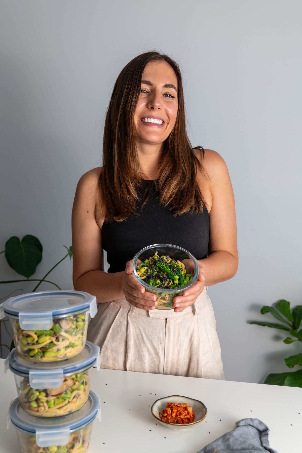Sarah Cobacho holding a glass container filled with high-protein udon noodle meal prep, smiling at the camera.
