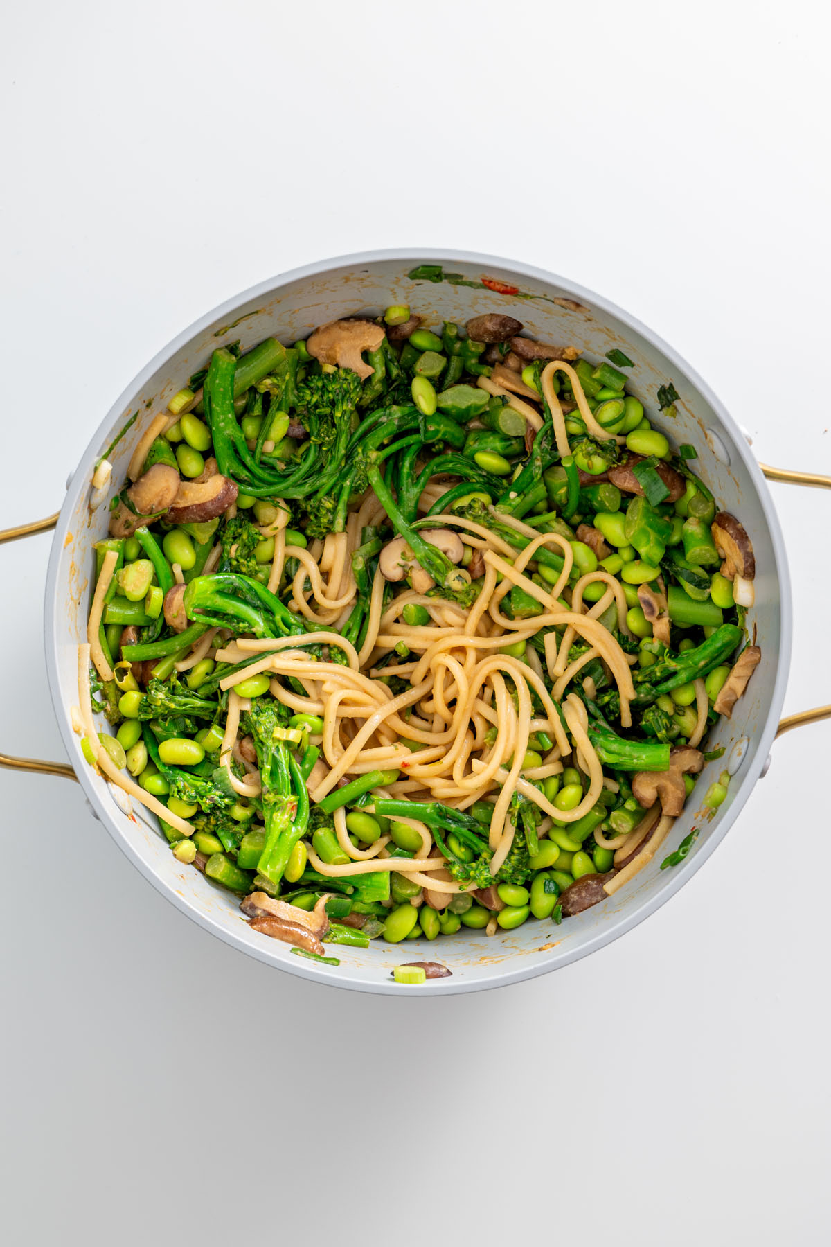 Close-up of a pot filled with high-protein udon noodles, broccolini, shiitake mushrooms, and edamame on a white surface.