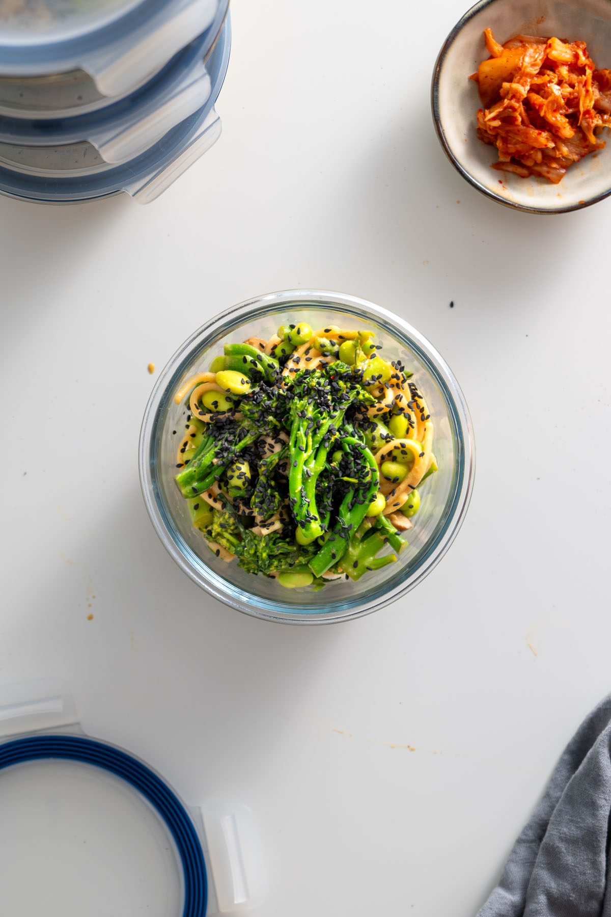 Glass container of high-protein udon noodles topped with black sesame seeds, surrounded by additional meal prep containers and a small dish of kimchi.