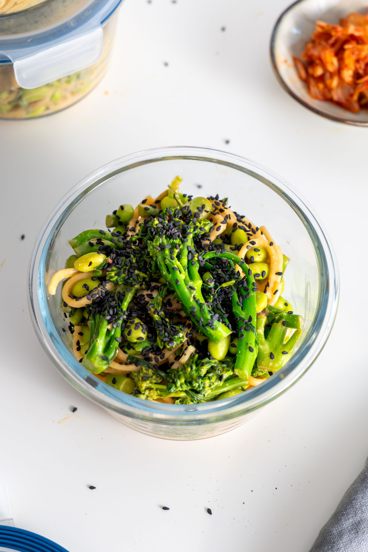 Close-up of high-protein udon noodles with broccolini and black sesame seeds in a glass container, placed on a white surface.