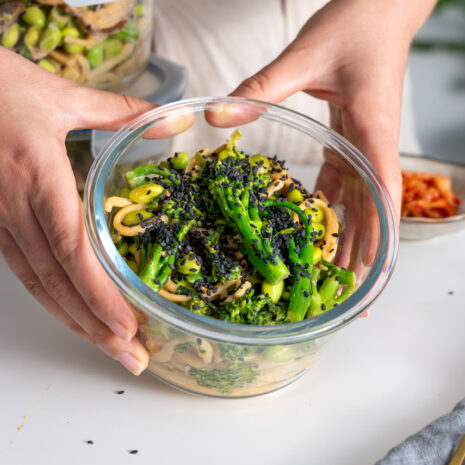 Sarah Cobacho holding a glass container of high-protein udon noodles, with additional meal prep containers visible in the background.