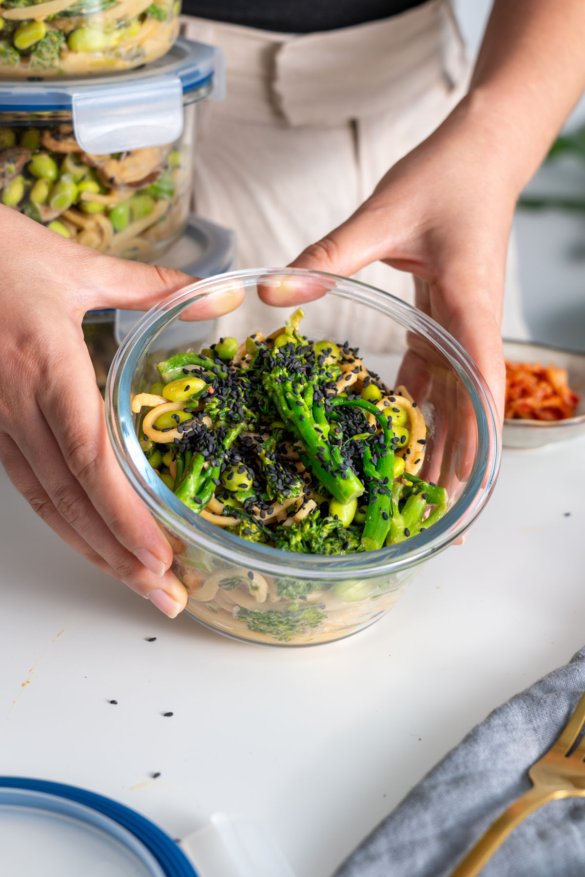 Sarah Cobacho holding a glass container of high-protein udon noodles, with additional meal prep containers visible in the background.