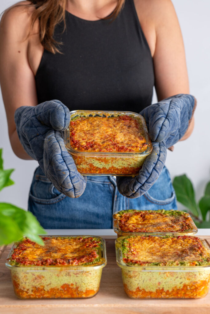 Sarah Cobacho holding a glass meal prep container filled with golden baked high-protein vegan lasagna.