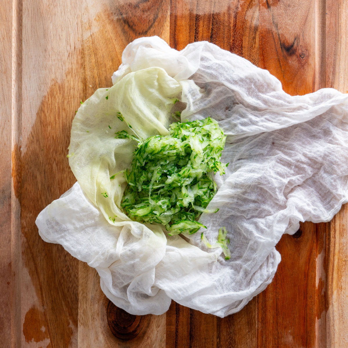 Grated cucumber being squeezed in a cheesecloth on a wooden cutting board.