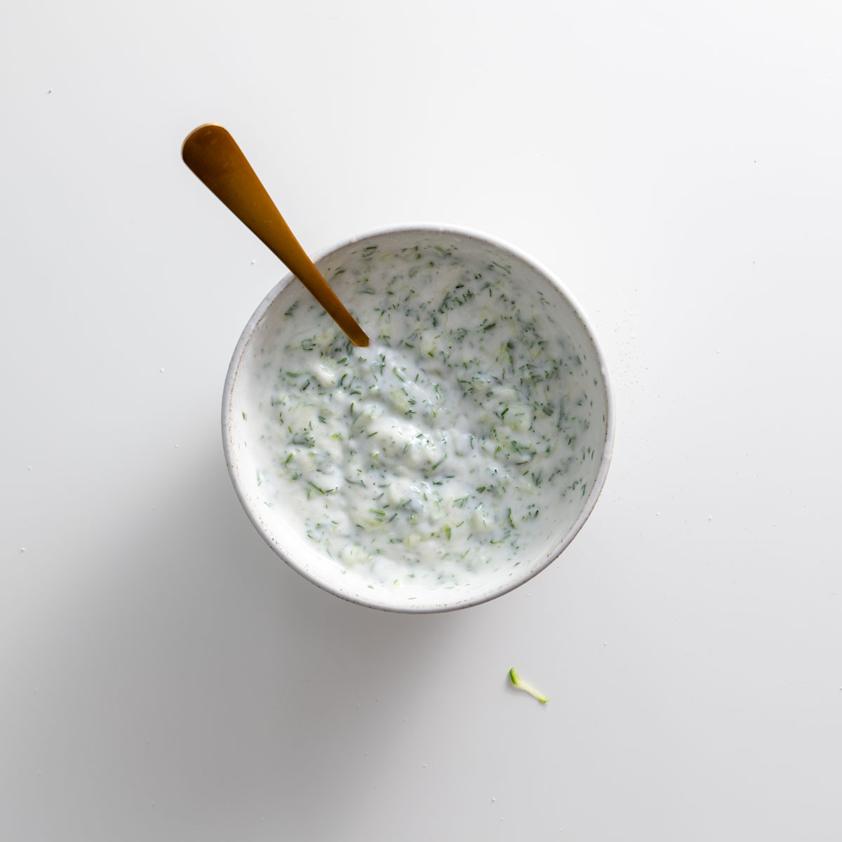 Prepared tzatziki in a white bowl with a spoon on a white background.
