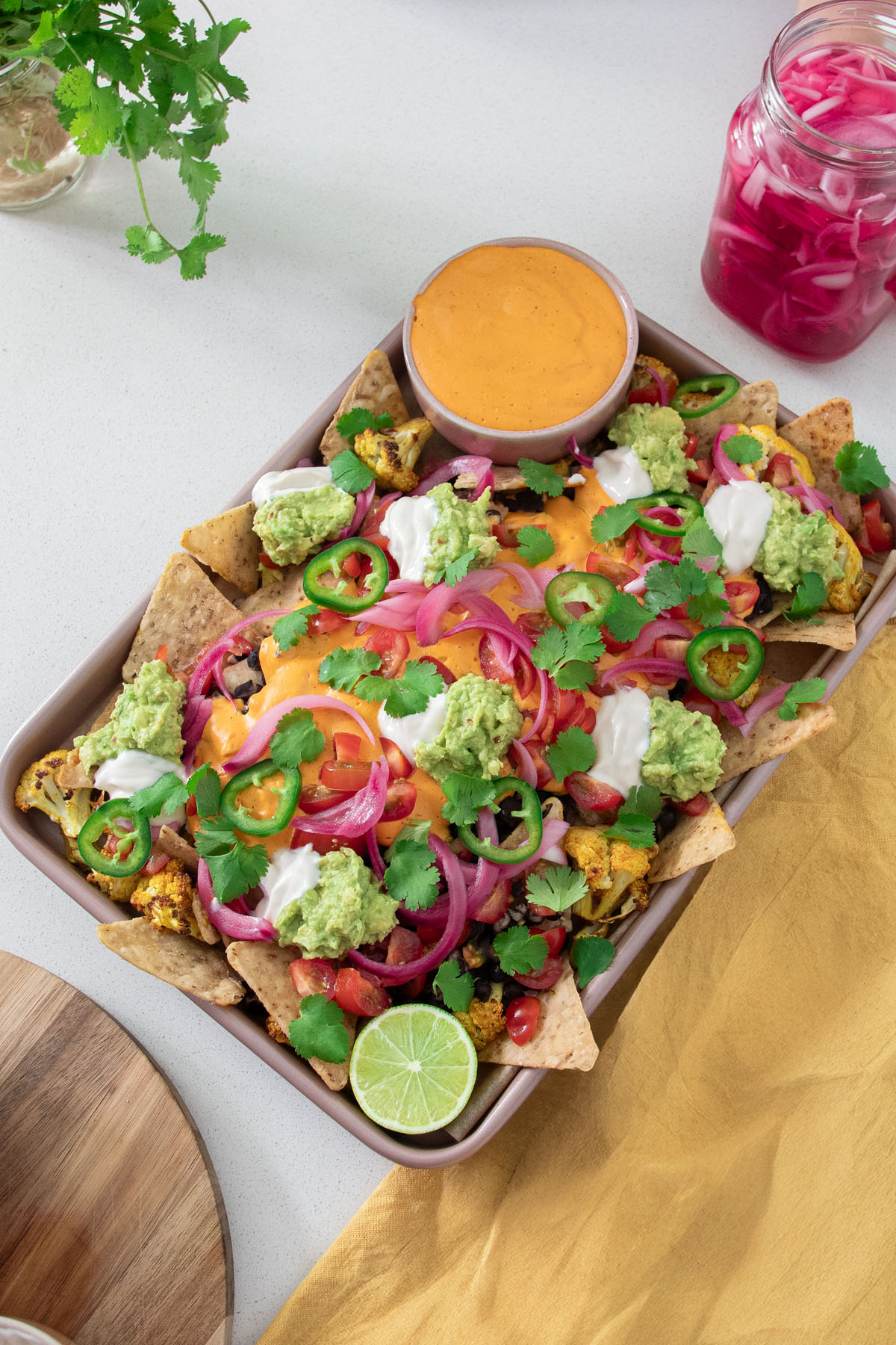 A vibrant tray of cauliflower nachos topped with dairy-free queso, guacamole, pickled onions, jalapeños, and fresh cilantro on a white countertop.