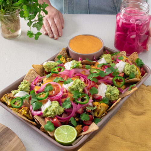 Sarah Cobacho standing behind a tray of colorful cauliflower nachos, with a jar of pickled onions and fresh cilantro nearby.