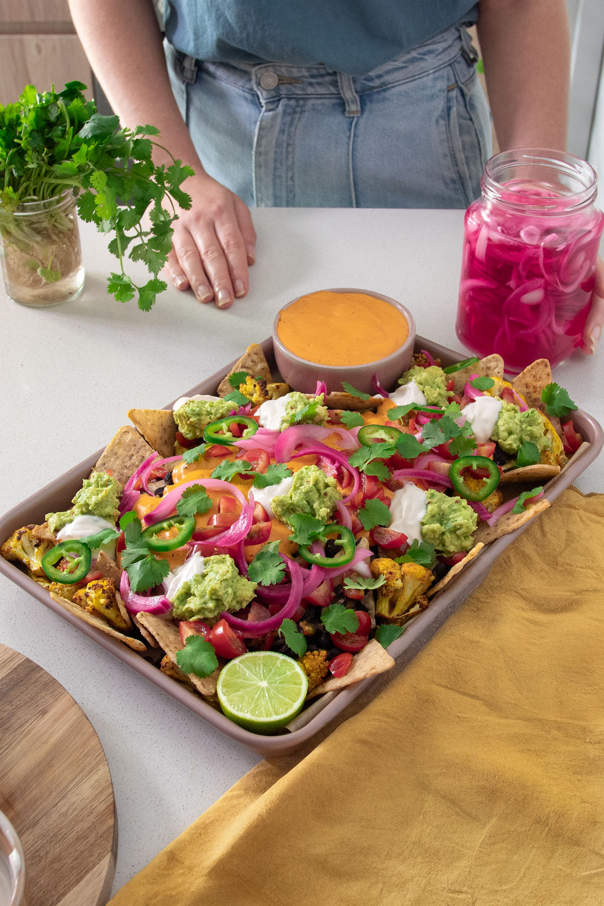 Sarah Cobacho standing behind a tray of colorful cauliflower nachos, with a jar of pickled onions and fresh cilantro nearby.
