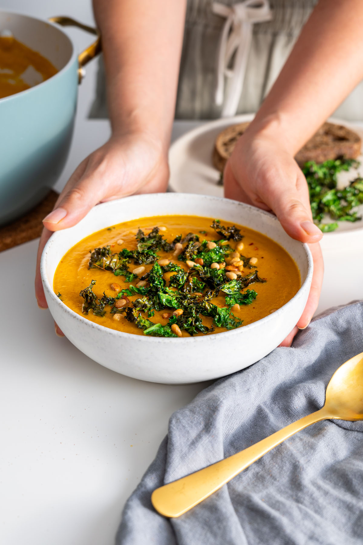 Sarah Cobacho holding a bowl of vibrant roasted vegetable soup topped with crispy kale and pine nuts.