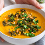 Close-up of roasted vegetable soup garnished with crispy kale and pine nuts.