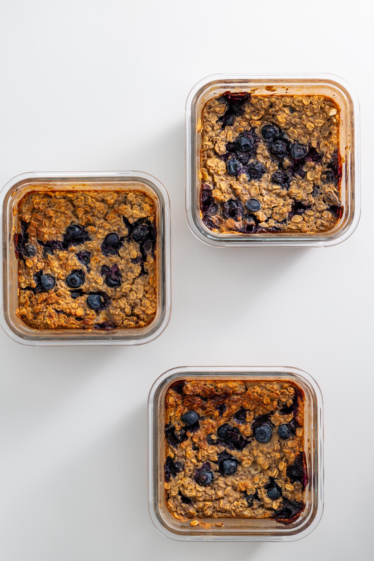 Three glass containers of baked blueberry oats, golden brown with blueberries on top.