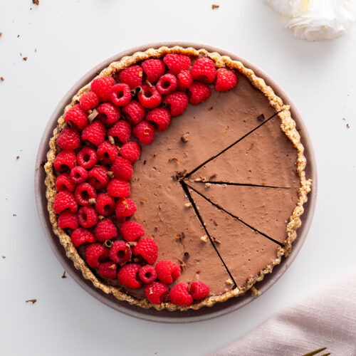 A no-bake silken chocolate tart with a nutty crust, decorated with fresh raspberries in a half-circle, partially sliced.