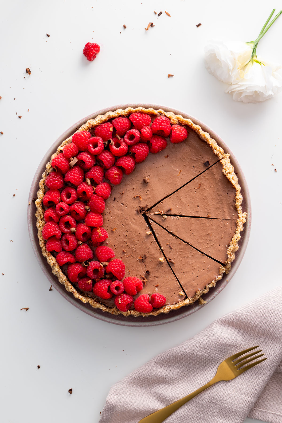 A no-bake silken chocolate tart with a nutty crust, decorated with fresh raspberries in a half-circle, partially sliced.