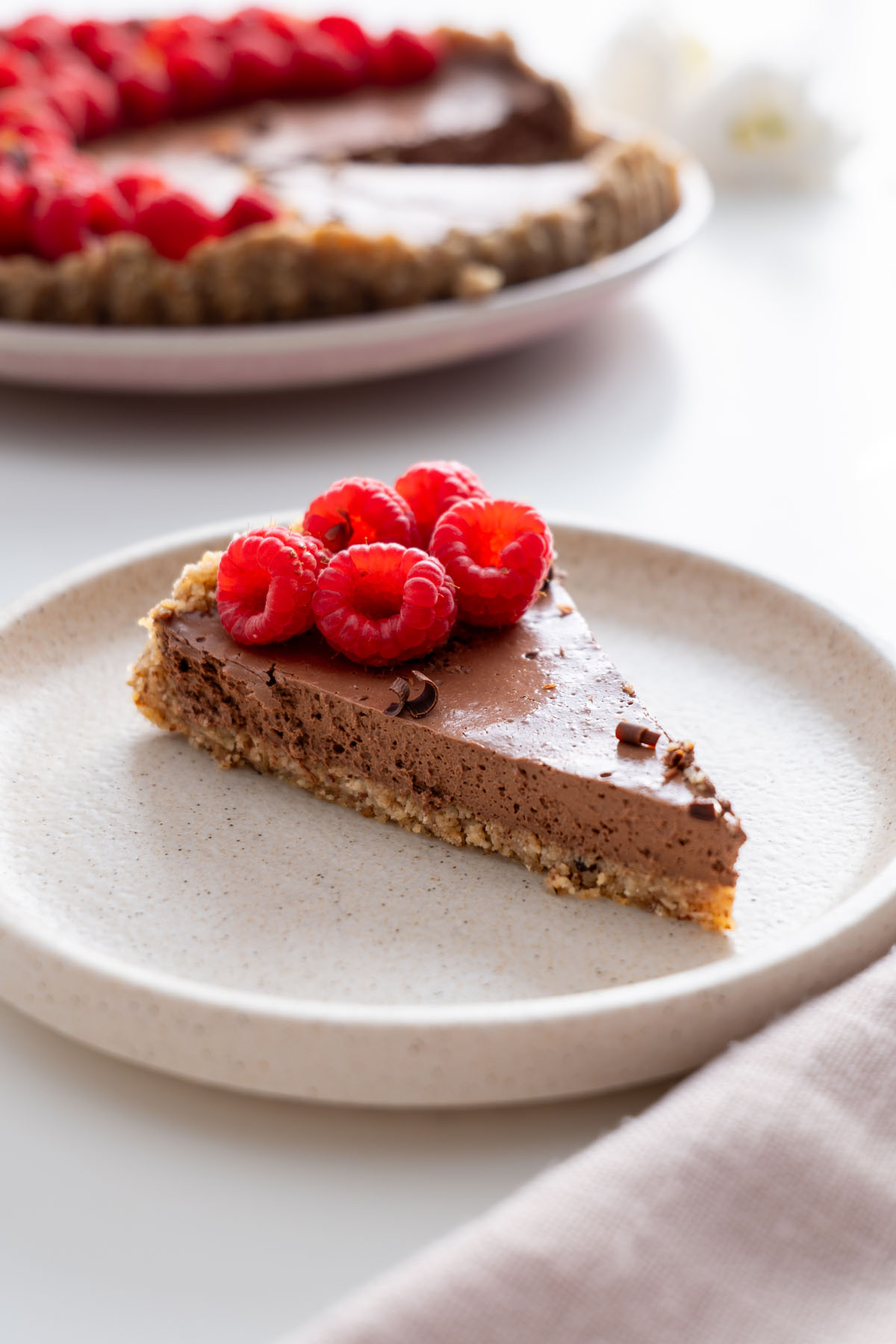A close-up of a single slice of no-bake chocolate tart with a smooth, creamy texture, topped with raspberries, with the whole tart in the background.