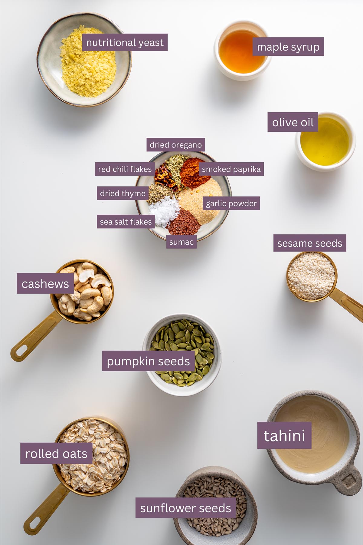 Ingredients for homemade savory granola, including tahini, nuts, seeds, and spices, arranged on a white surface.