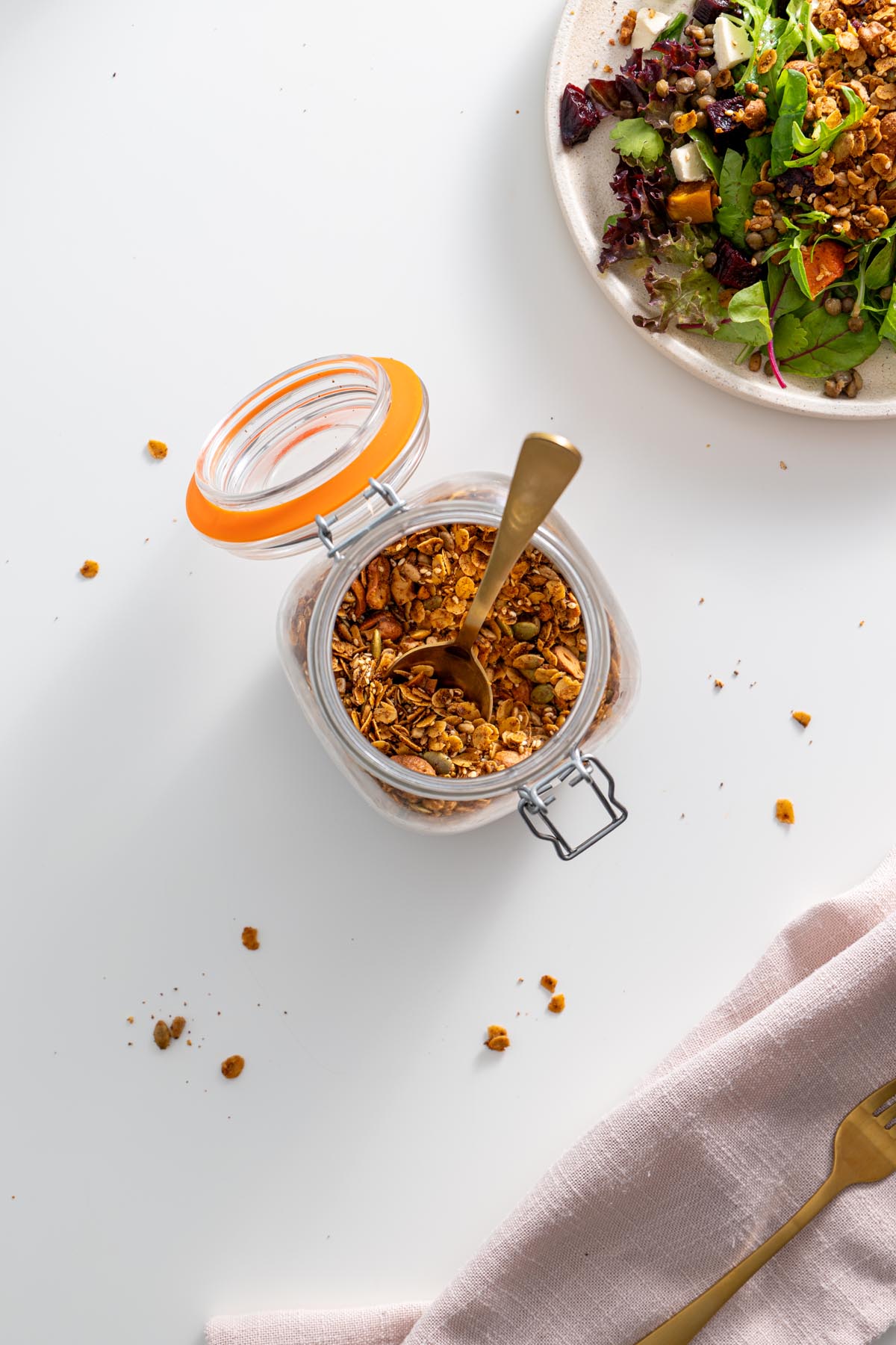 Jar of homemade savory granola with a gold spoon, placed on a white surface with a salad in the background.