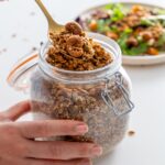Sarah Cobacho holding a gold spoon scooping homemade savory granola from a glass jar.