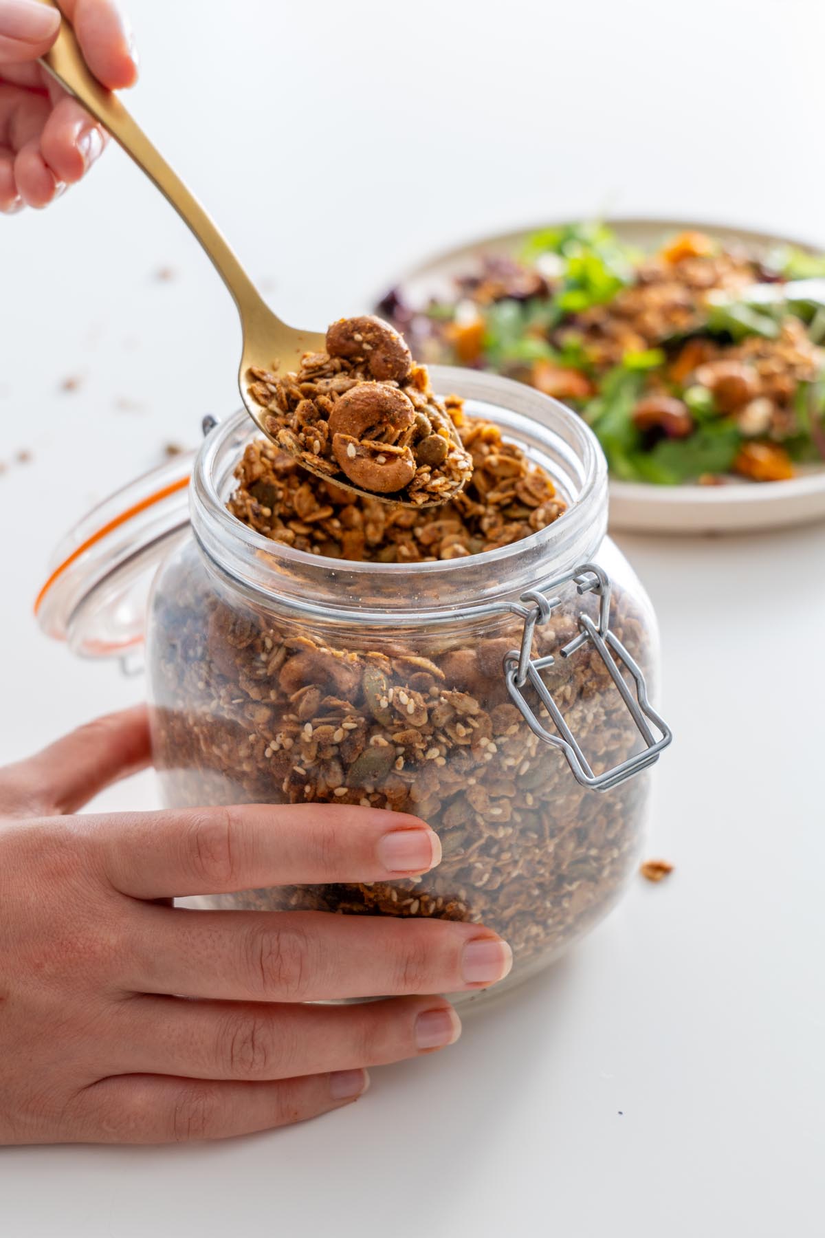 Sarah Cobacho holding a gold spoon scooping homemade savory granola from a glass jar.