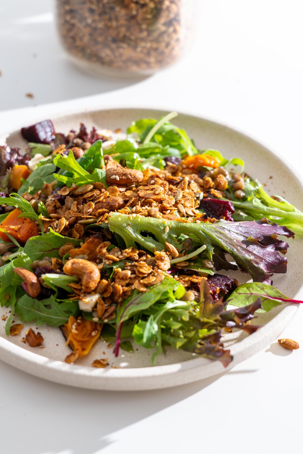 Close-up of a salad topped with crunchy homemade savory granola, with a jar of granola in the background.