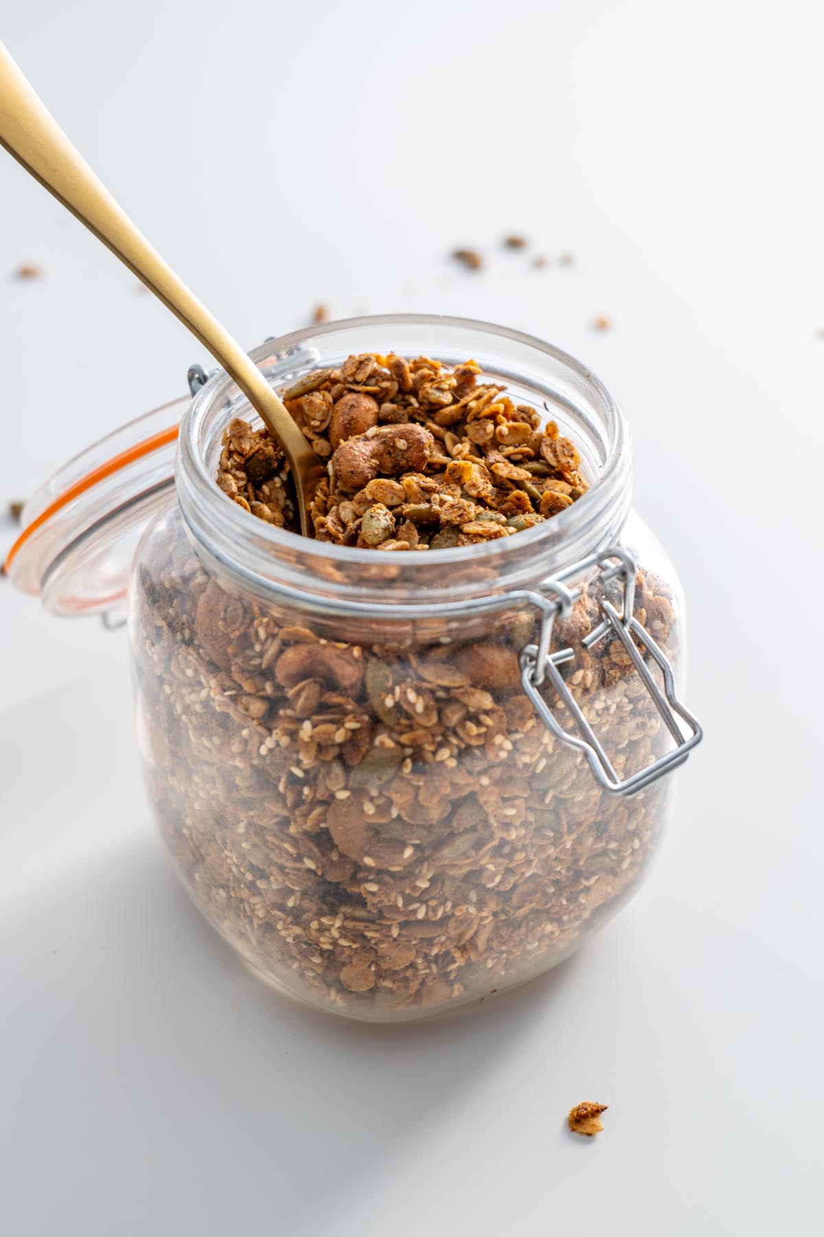 A jar of freshly baked savory granola with a gold spoon resting inside, placed on a clean white surface.
