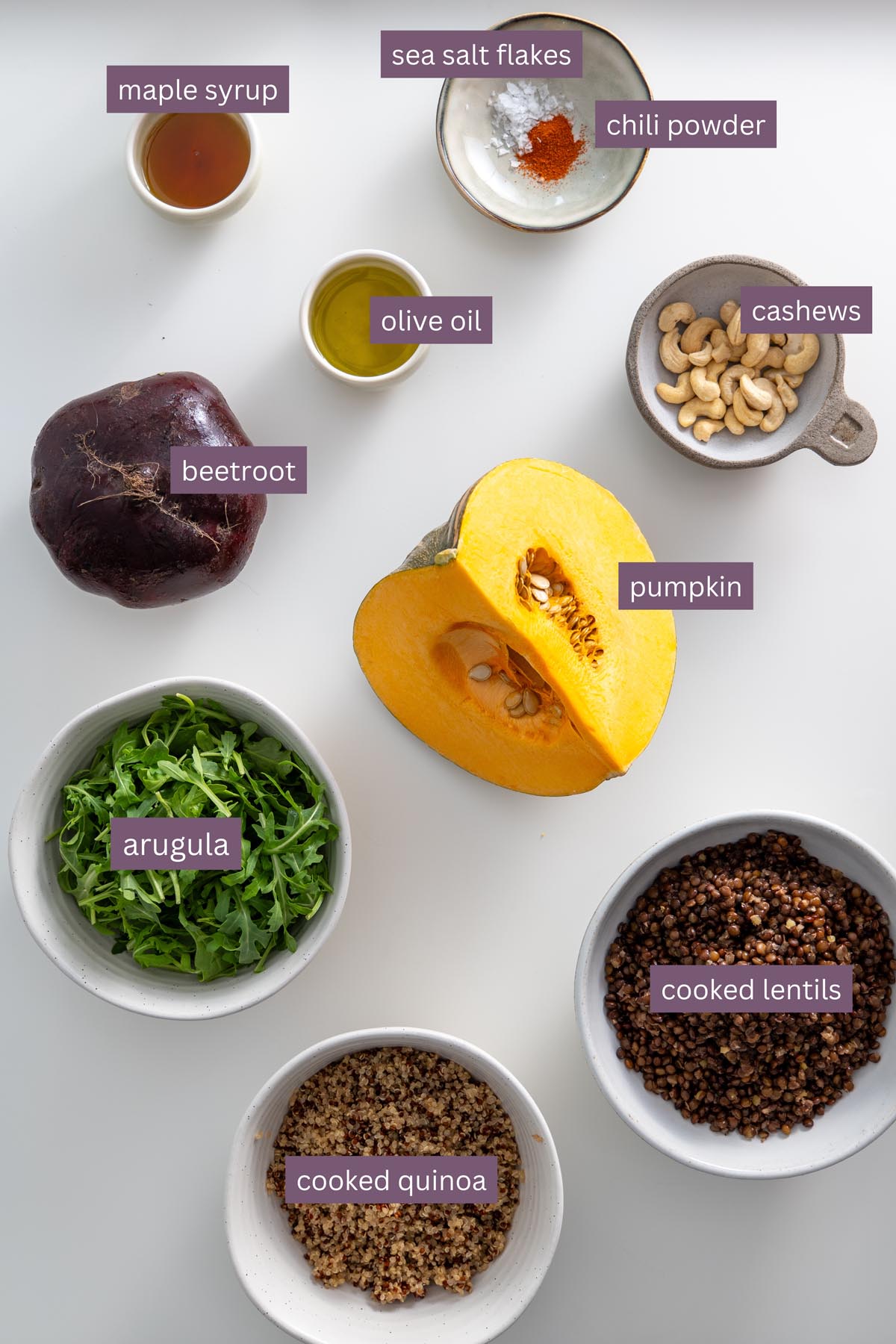 Vibrant ingredients for a dill pickle salad on a white surface, including beetroot, pumpkin, quinoa, lentils, cashews, and arugula.