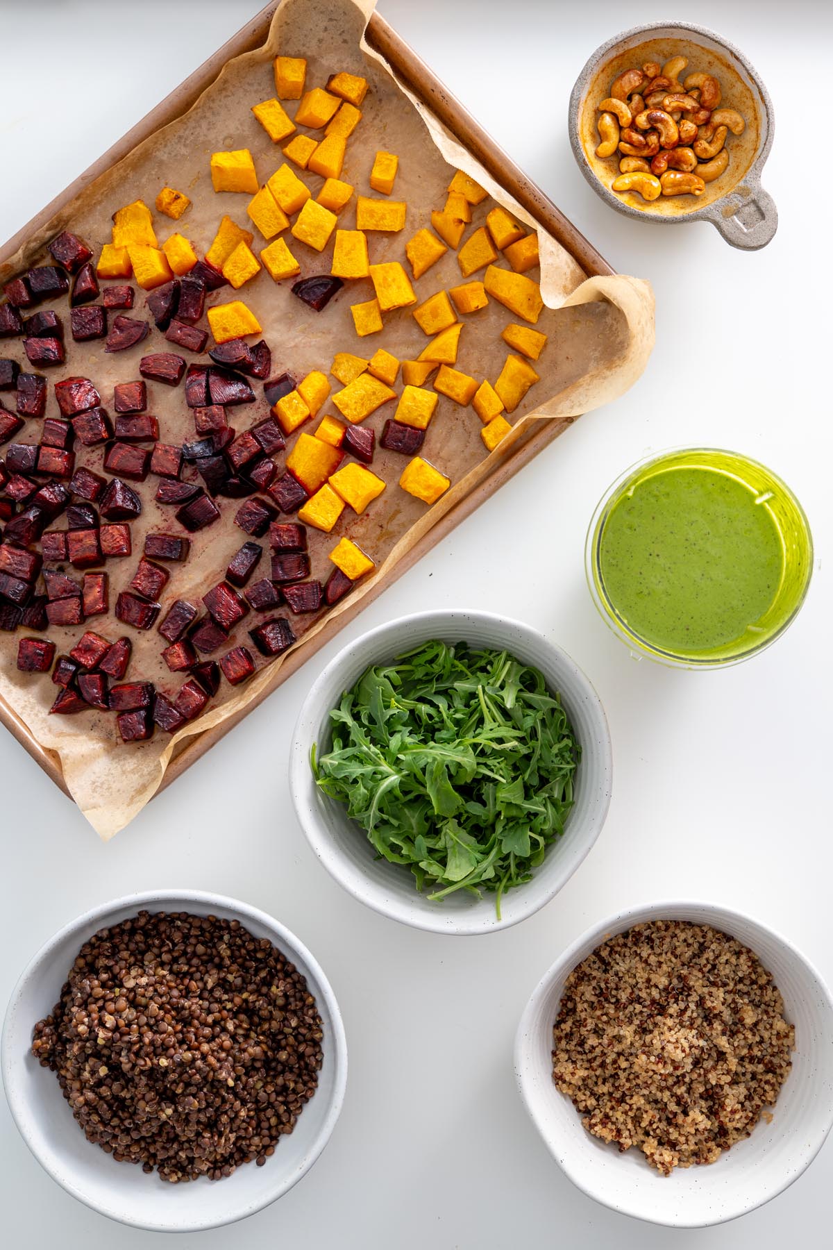 Fully prepared dill pickle salad ingredients arranged on a white surface, including roasted vegetables, quinoa, lentils, arugula, and dressing.