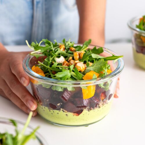 Sarah Cobacho holding a glass meal prep container filled with dill pickle salad, showcasing layers of roasted vegetables, quinoa, and dressing.