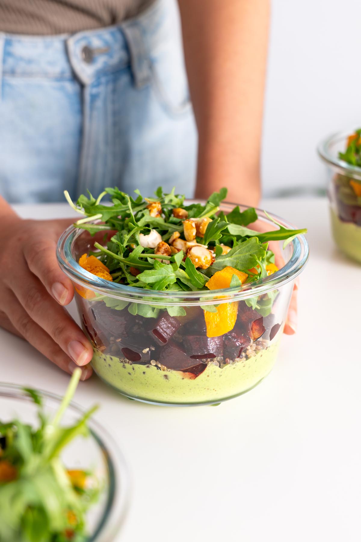 Sarah Cobacho holding a glass meal prep container filled with dill pickle salad, showcasing layers of roasted vegetables, quinoa, and dressing.