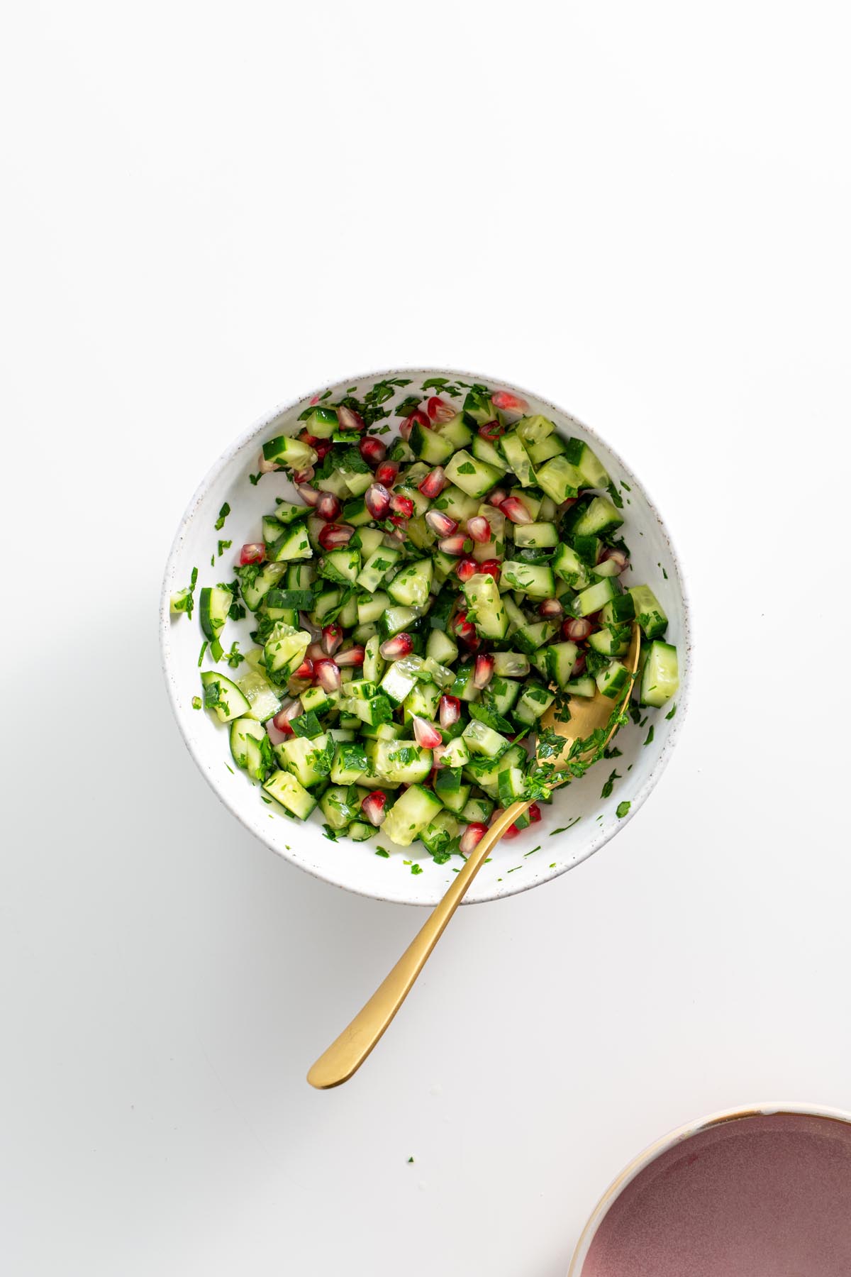 Fresh cucumber and pomegranate salad in a white bowl with a gold spoon.