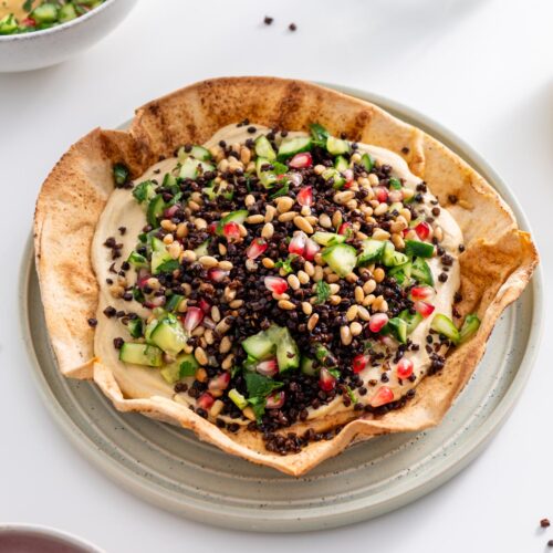 Freshly baked pita bowl filled with hummus, topped with cucumber salad, lentils, and pine nuts.