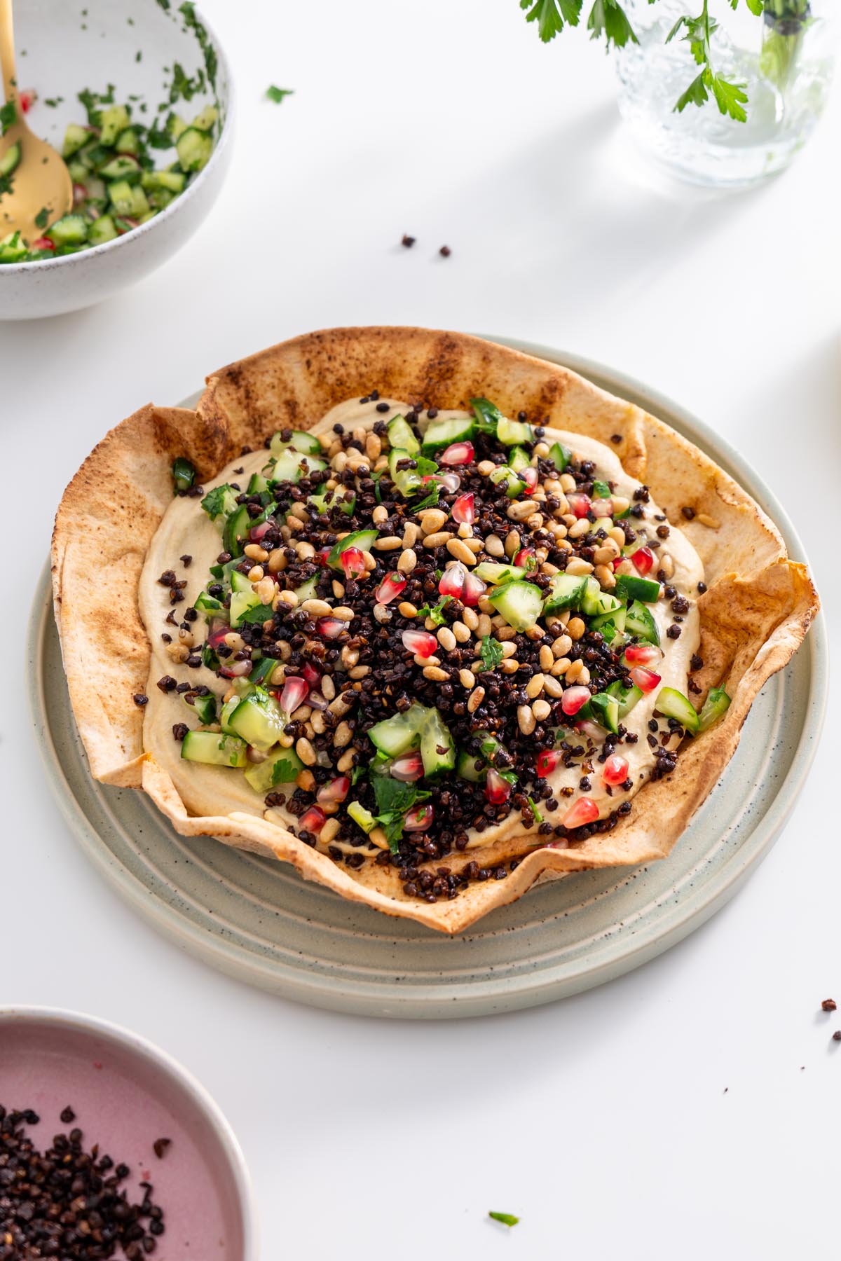 Freshly baked pita bowl filled with hummus, topped with cucumber salad, lentils, and pine nuts.