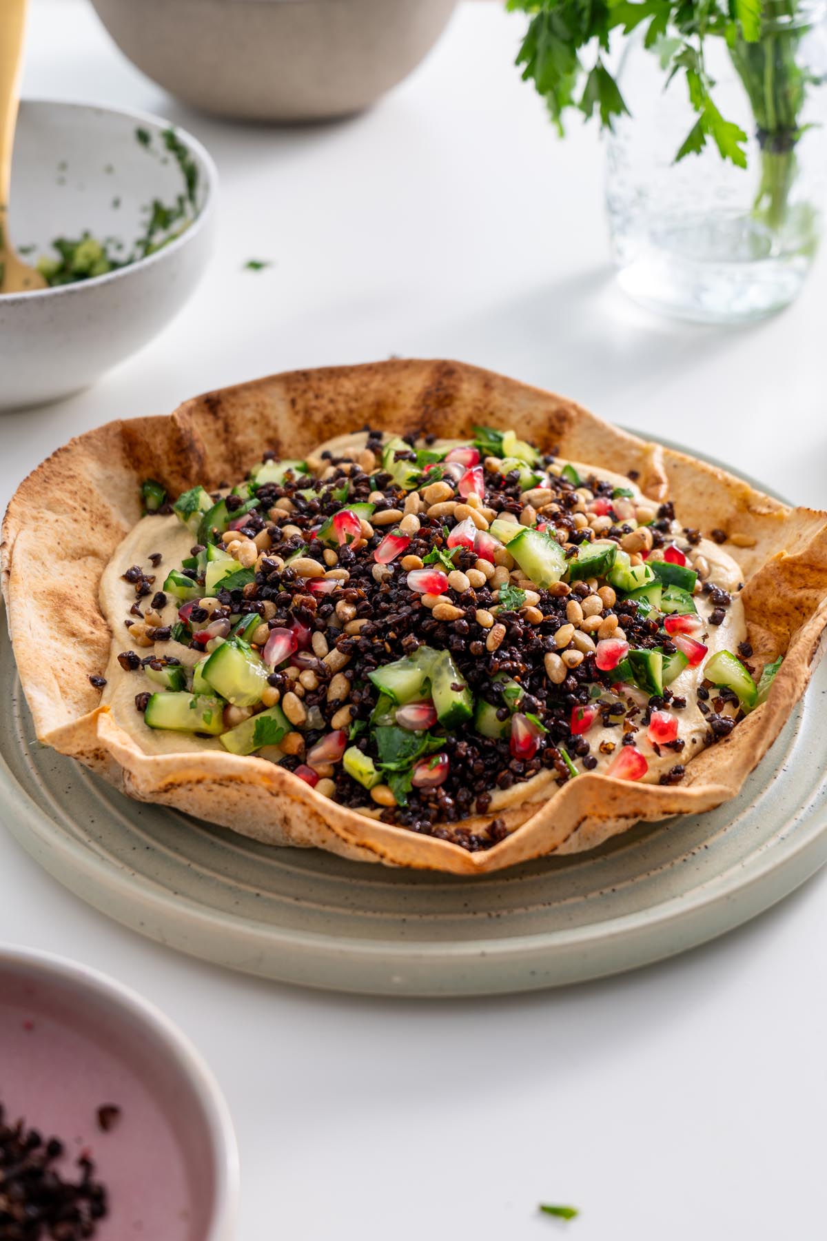 Freshly baked pita bowl filled with hummus, topped with cucumber salad, lentils, and pine nuts.