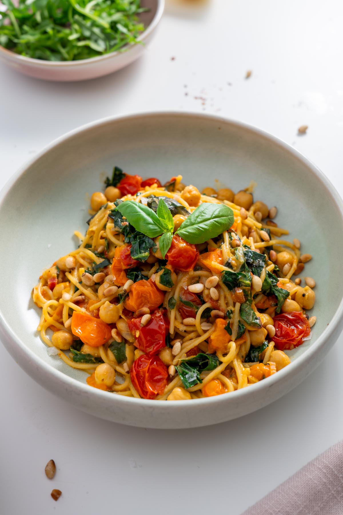 A bowl of Meghan Markle’s one-pot pasta with cherry tomatoes, chickpeas, kale, and fresh basil on top, served on a white table.