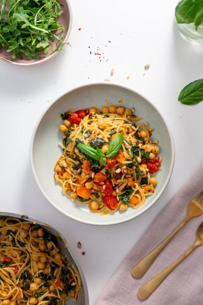 A plated bowl of Meghan Markle’s one-pot pasta on a white table with fresh arugula, chili flakes, and gold cutlery in the background.