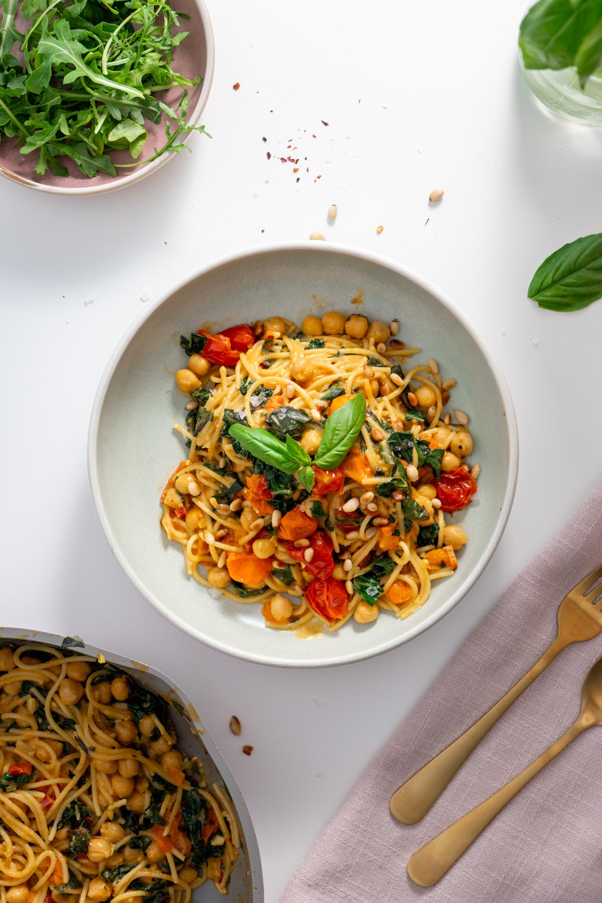 A plated bowl of Meghan Markle’s one-pot pasta on a white table with fresh arugula, chili flakes, and gold cutlery in the background.