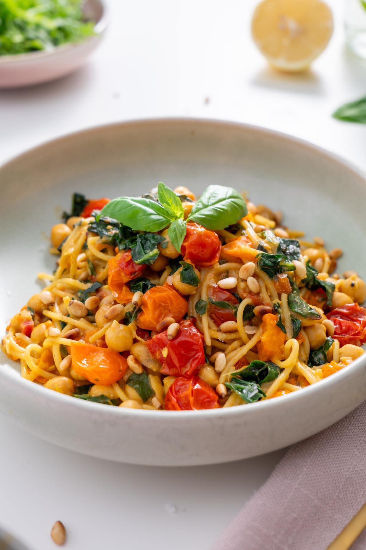Close-up of a bowl of Meghan Markle’s one-pot pasta with bright cherry tomatoes, chickpeas, kale, and toasted pine nuts, garnished with fresh basil.
