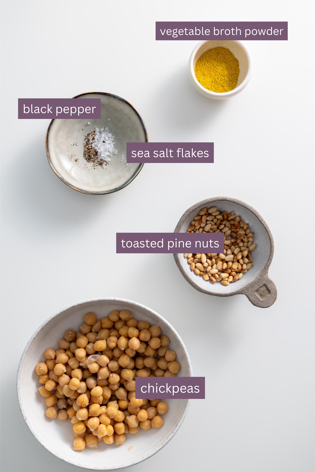 A flat lay of key ingredients for the Meghan Markle one-pot pasta, including chickpeas, pine nuts, sea salt, black pepper, and vegan parmesan on a white surface.