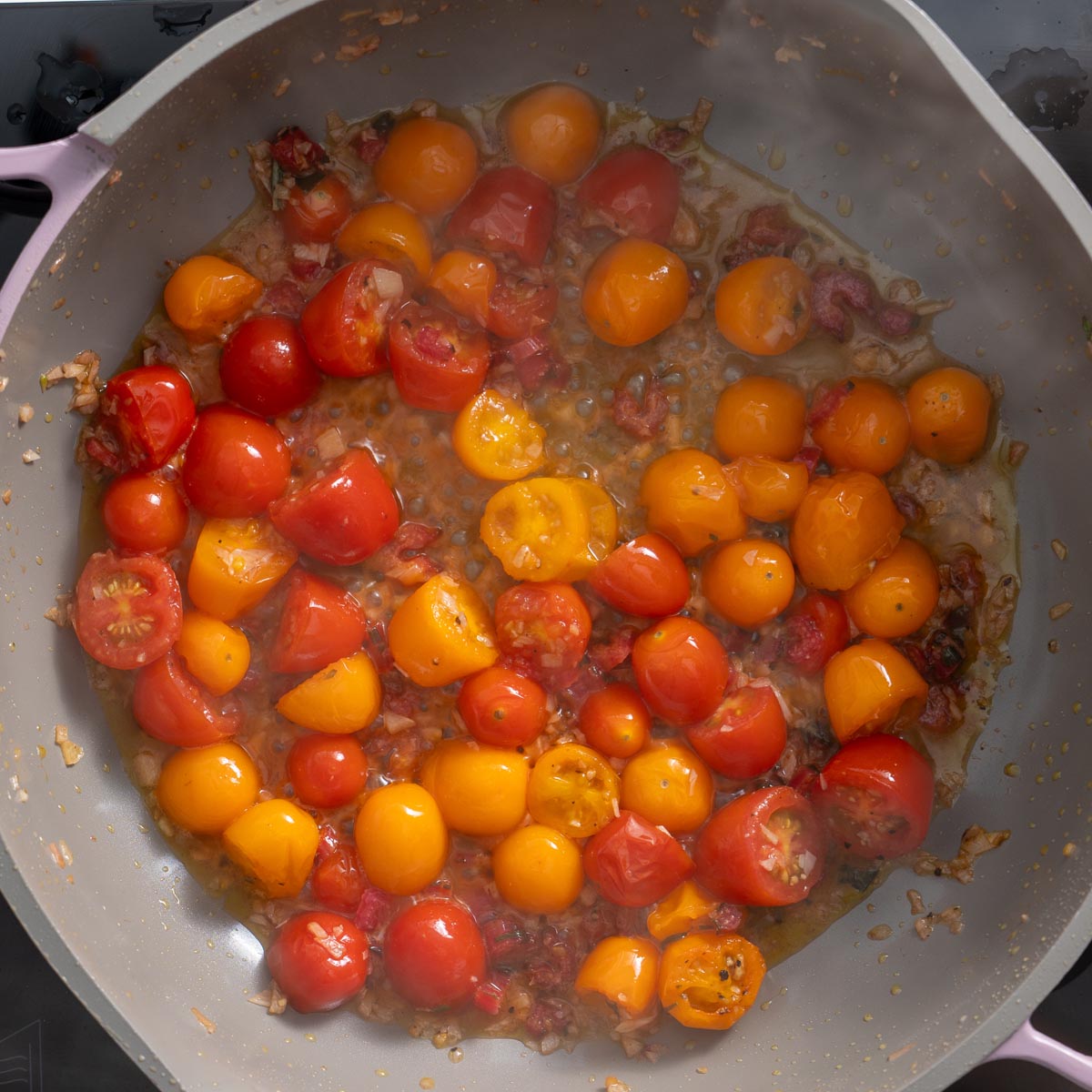 Golden cherry tomatoes and red tomatoes sizzling in a pan with garlic and seasonings, releasing their juices.