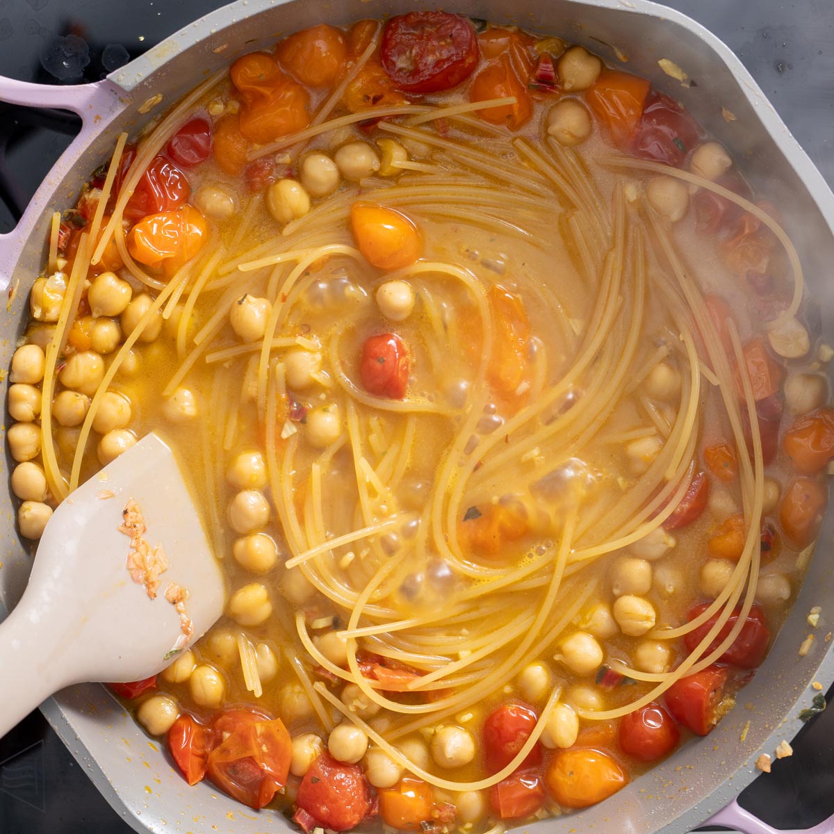 Spaghetti and chickpeas cooking in a golden broth with softened tomatoes and seasonings in a large pan.