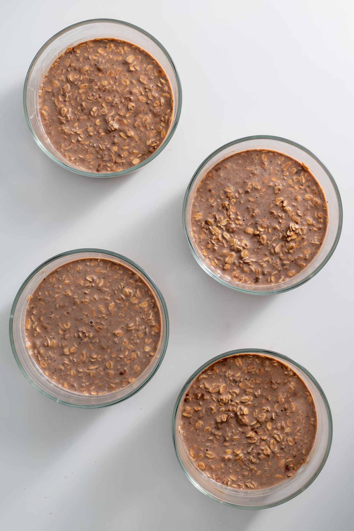 Four glass bowls filled with unbaked tiramisu oat mixture on a white surface
