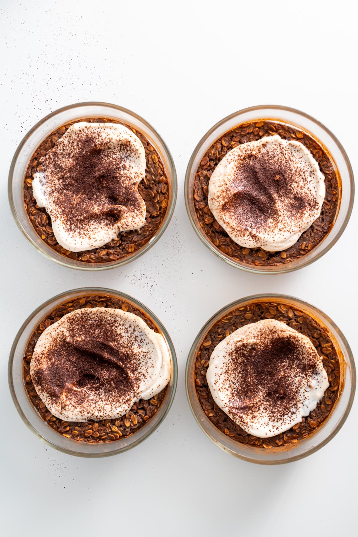 Four servings of tiramisu baked oats, topped with yogurt and cacao powder, arranged on a white surface