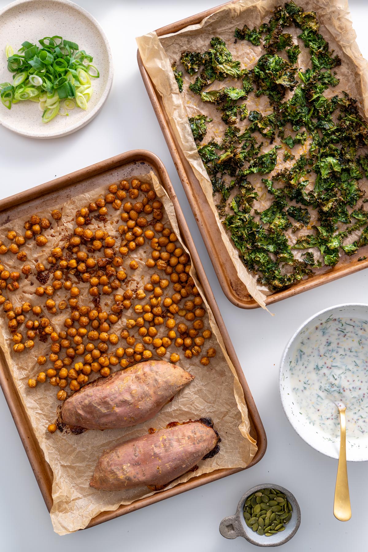 Sliced scallions, crispy kale, roasted chickpeas, and sweet potatoes on trays with sauce and seeds.
