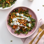 Two bowls of kale sweet potato salad topped with chickpeas and yogurt sauce on a white table.