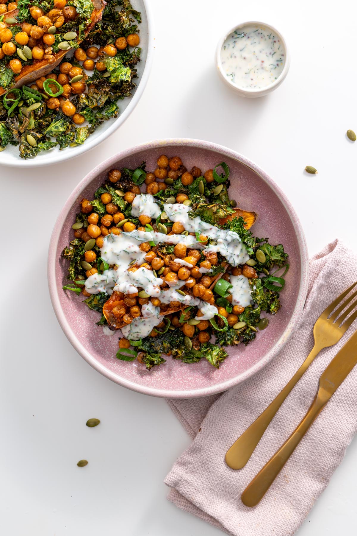 Two bowls of kale sweet potato salad topped with chickpeas and yogurt sauce on a white table.