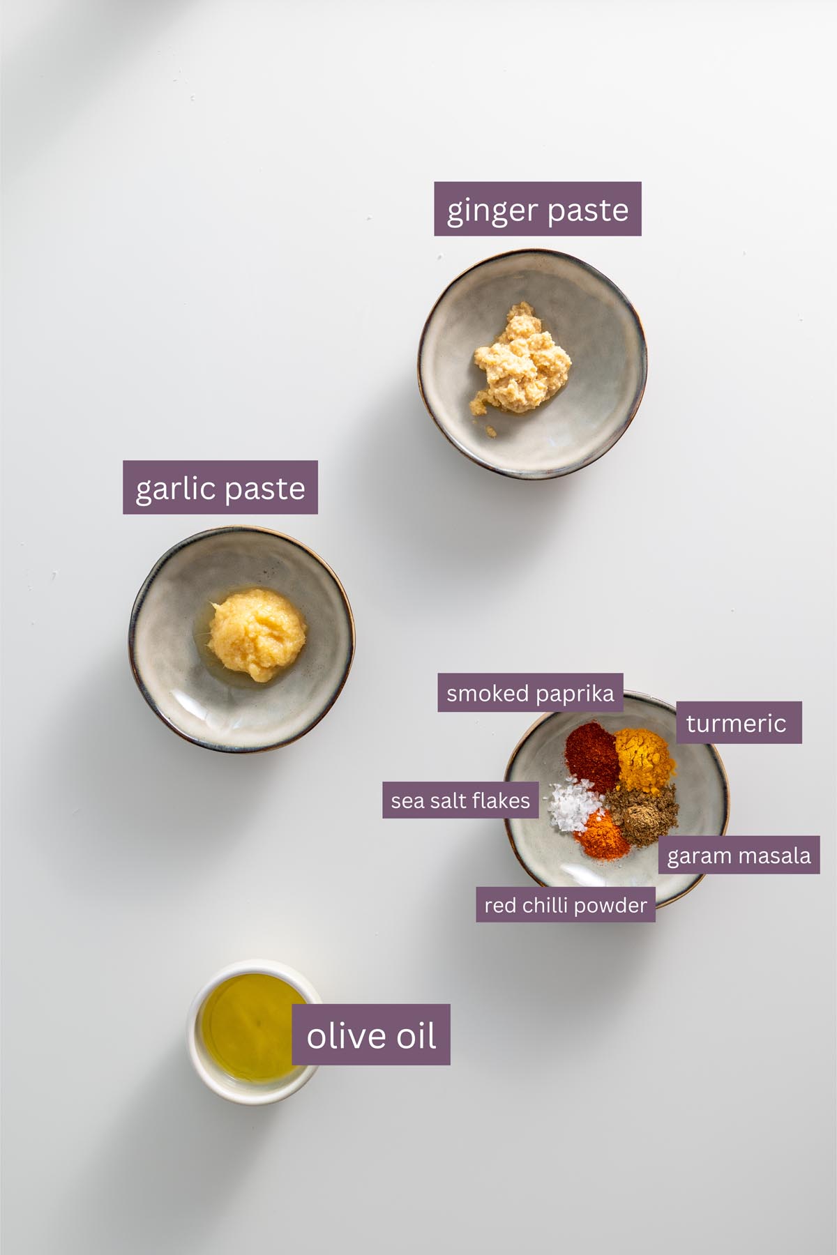 A variety of tandoori spices, garlic paste, and ginger paste in small bowls on a white surface.