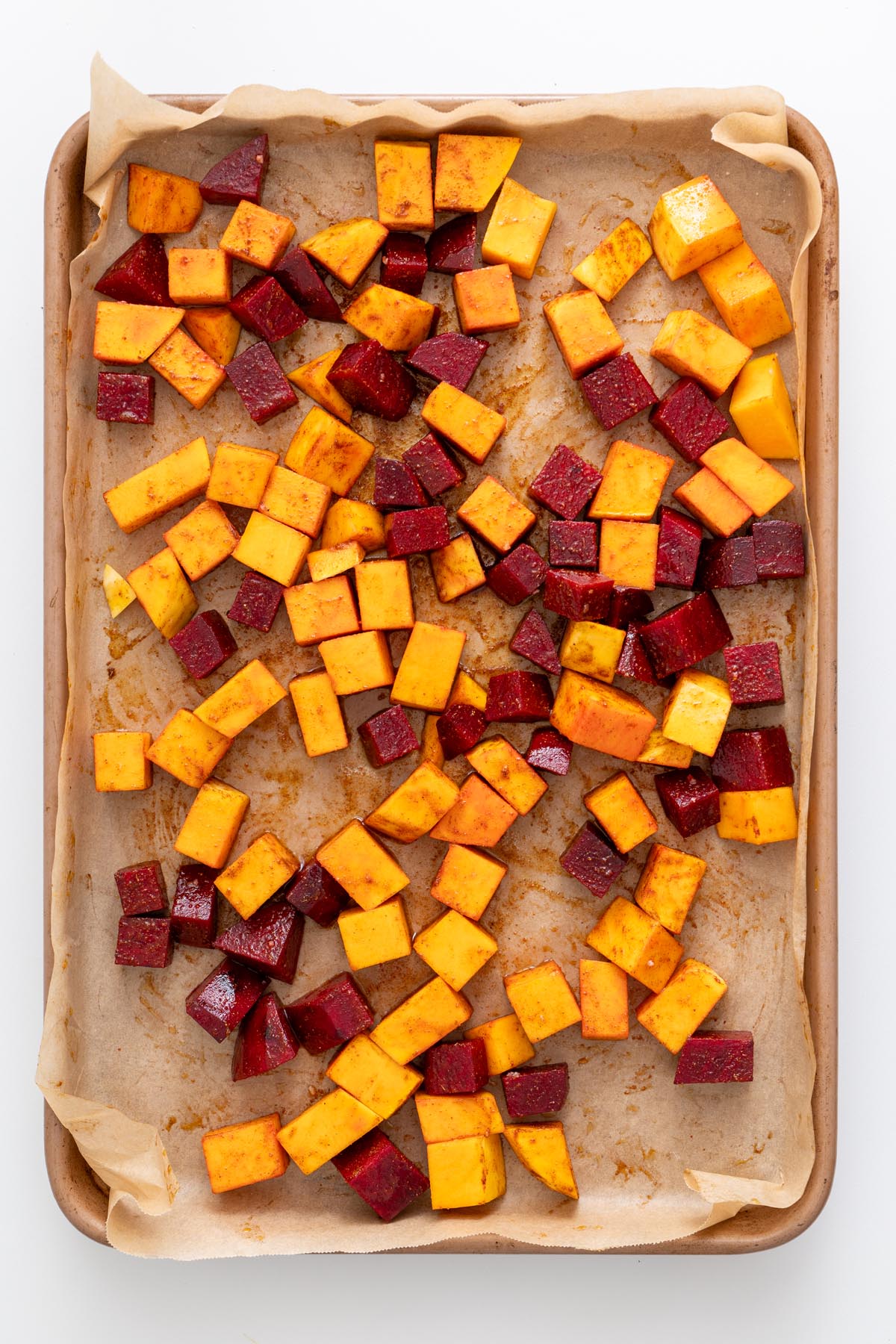 Diced raw butternut squash and beet tossed with spices on a baking tray lined with parchment paper.