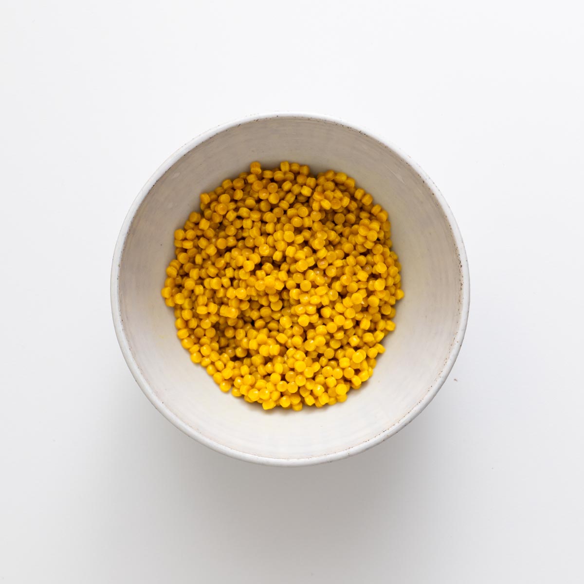 Cooked turmeric pearl couscous in a small white bowl on a white background.